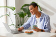 © Prostock-studio - Smiling young latin lady enjoy cup of coffee and croissant at table and video call on laptop