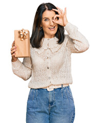 Poster - Middle age brunette woman holding gift smiling happy doing ok sign with hand on eye looking through fingers