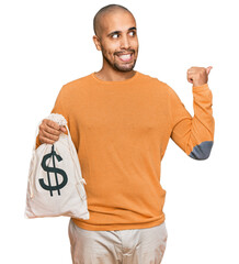 Sticker - Hispanic adult man holding dollars bag pointing thumb up to the side smiling happy with open mouth