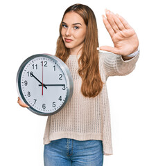 Poster - Young blonde woman holding big clock with open hand doing stop sign with serious and confident expression, defense gesture