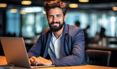 Wall Mural - Confident and cheerful young professional man with a beard sitting at his desk with a laptop in a modern office space, looking at the camera with a friendly smile