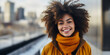 Radiant young African American woman with afro hair smiling brightly, dressed in winter fashion, on a city bridge with blurred urban skyline in the background