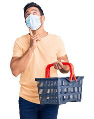 Wall Mural - Young hispanic man wearing shopping basket and medical mask serious face thinking about question with hand on chin, thoughtful about confusing idea