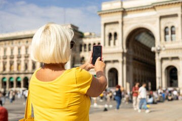 Beautiful senior blonde woman taking selfie on Duomo square, Galleria Vittorio Emanuele II shopping arcade in Milan, Italy. Traveling Europe in summer. Attractive 60s female is exploring city