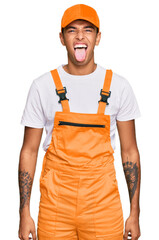 Canvas Print - Young handsome african american man wearing handyman uniform sticking tongue out happy with funny expression. emotion concept.