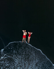 Sticker - Black sand beach. Fantastic landscape of black sand volcano beach. Couple having a good time on vacation. A man and a woman in red swimsuits are lying down