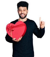 Sticker - Young arab man with beard holding valentine gift smiling with an idea or question pointing finger with happy face, number one