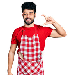 Poster - Young arab man with beard wearing cook apron smiling and confident gesturing with hand doing small size sign with fingers looking and the camera. measure concept.