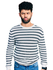 Sticker - Young arab man with beard wearing casual striped sweater making fish face with lips, crazy and comical gesture. funny expression.