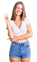 Wall Mural - Young beautiful blonde woman wearing casual white tshirt smiling with happy face winking at the camera doing victory sign with fingers. number two.