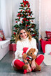 Cheerful pretty mom in white knitted sweater and red leggings with a Christmas teddy bear toy against the background of the Christmas tree (New Year, 2024, Christmas - concept)