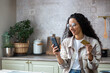 © Liubomir - Young beautiful woman in kitchen with phone and bank credit debit card in hands, smiling Hispanic woman chooses gifts and products in online store, online shopping remotely from home.