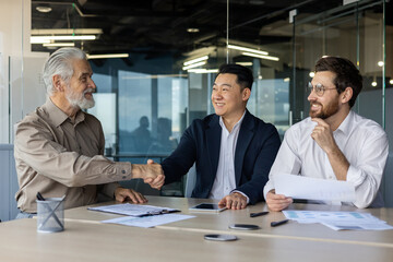 Making a business deal in the office. Three interracial men of different ages sit at a table with documents and negotiate while shaking hands.