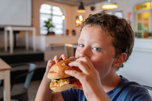 Portrait Of Hungry Cute Elementary Boy Eating Delicious Burger And Looking At Camera While Spending Leisure Time In Restaurant At Weekend