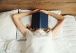 Young sad tired woman student covering her face with open book with blank cover while lying on the bed at home