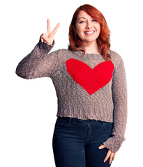 Wall Mural - Young beautiful redhead woman wearing casual heart sweater smiling with happy face winking at the camera doing victory sign. number two.