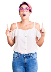 Canvas Print - Young beautiful woman with pink hair wearing casual clothes and glasses amazed and surprised looking up and pointing with fingers and raised arms.