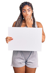 Canvas Print - Young african american woman with braids holding blank empty banner covering mouth with hand, shocked and afraid for mistake. surprised expression