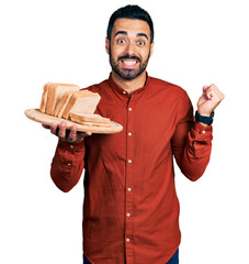 Wall Mural - Young hispanic man with beard holding bread screaming proud, celebrating victory and success very excited with raised arm