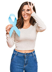 Wall Mural - Young brunette woman holding blue ribbon smiling happy doing ok sign with hand on eye looking through fingers