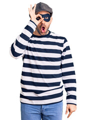 Wall Mural - Young handsome man wearing burglar mask doing ok gesture shocked with surprised face, eye looking through fingers. unbelieving expression.