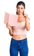 Canvas Print - Young beautiful woman holding book pointing with finger to the camera and to you, confident gesture looking serious