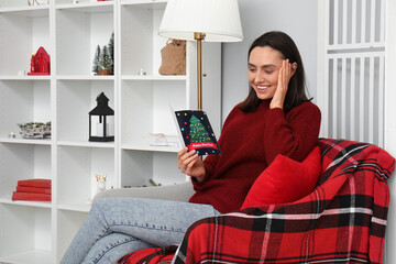 Canvas Print - Happy young woman reading Christmas greeting card while sitting on armchair at home