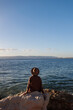 © zhukovvvlad - Back view. relaxed girl in a hat and coat sitting in front of the sea on the stones.