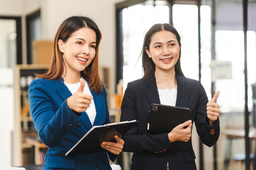 Two middle aged and young Asian female executives in formal suits review bar chart, discussing business strategies in office setting, senior executives or directors in advertising or public relations