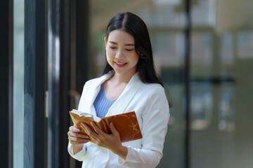 A pretty and confident Asian businesswoman standing and reading a book in an office.