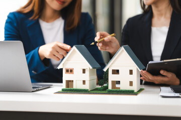 Two Asian real estate agents in formal suits discuss a contract with house models on the table, ready to sign a deal.