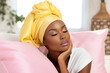 Portrait of a beautiful young African woman in headscarf sitting on a sofa in a living room in light pastel colored interior.