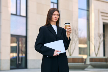 Portrait of a young woman in a business suit drinking coffee and holding a laptop outside. A confident and successful woman