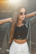 © alones - Beautiful fashionable fresh young girl with vintage sunglasses in a fashion outfit with a shirt straightens her hair near a black brick wall at sunset. Pretty lady with sunlight