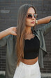 © alones - Beautiful glamorous young stylish woman model with sunglasses in fashion clothes with a green shirt with a black top stands near a black brick wall. Pretty chic street girl at sunset