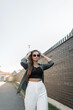 © alones - Beautiful fashionable street girl model with sunglasses in a fashion outfit with a shirt walks in the city near a black brick wall