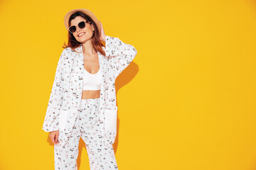 Wall Mural - Young beautiful smiling female in trendy summer costume clothes. Carefree woman posing near yellow wall in studio. Positive model having fun indoors. Cheerful and happy. In hat and sunglasses