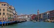 Bright sunny day at the Grand Canal with the Rialto Bridge in Venice, bustling with tourists and gondolas under a clear blue sky with fluffy clouds.