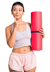 Sticker - Young woman holding yoga mat serious face thinking about question with hand on chin, thoughtful about confusing idea