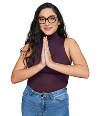 Wall Mural - Brunette young woman wearing casual clothes and glasses praying with hands together asking for forgiveness smiling confident.