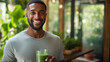 Cheerful young man with a distinctive hairstyle, holding a green smoothie, standing in front of a blurred background with indoor plants.