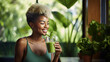 © MP Studio - Cheerful young woman with a distinctive hairstyle, holding a green smoothie, standing in front of a blurred background with indoor plants.