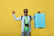 Happy young African man in sunglasses holding a large blue suitcase in his hand, pointing his finger up. Dark-skinned male traveler model demonstrates a roomy suitcase for trips on a yellow background
