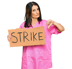 Canvas Print - Middle age brunette doctor woman holding strike banner cardboard with angry face, negative sign showing dislike with thumbs down, rejection concept