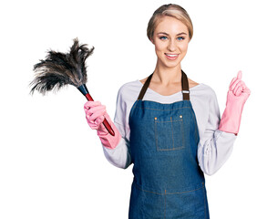 Poster - Beautiful young blonde woman wearing apron holding cleaning duster cheerful with a smile on face pointing with hand and finger up to the side with happy and natural expression