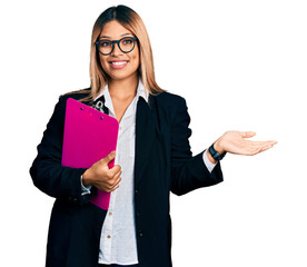 Young hispanic business woman holding clipboard with blank space smiling cheerful presenting and pointing with palm of hand looking at the camera.