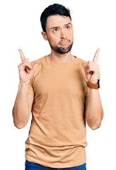 Poster - Hispanic man with beard wearing casual t shirt pointing up looking sad and upset, indicating direction with fingers, unhappy and depressed.