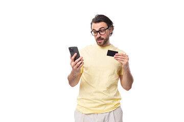 Wall Mural - casual caucasian guy with a beard in glasses dressed in a light t-shirt uses a smartphone and a plastic card