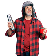 Canvas Print - Middle age hispanic man wearing hat with ear flaps drinking a cup of coffee holding thermo angry and mad screaming frustrated and furious, shouting with anger looking up.