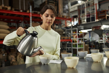 Wall Mural - Waist up portrait of young woman pouring hot water into small bowls during cupping and quality control inspection at coffee factory, copy space